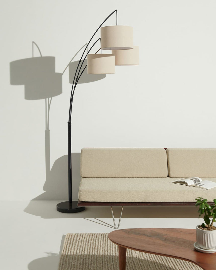 Black three arm lamp beside a cream sofa, a low wood table and a plant