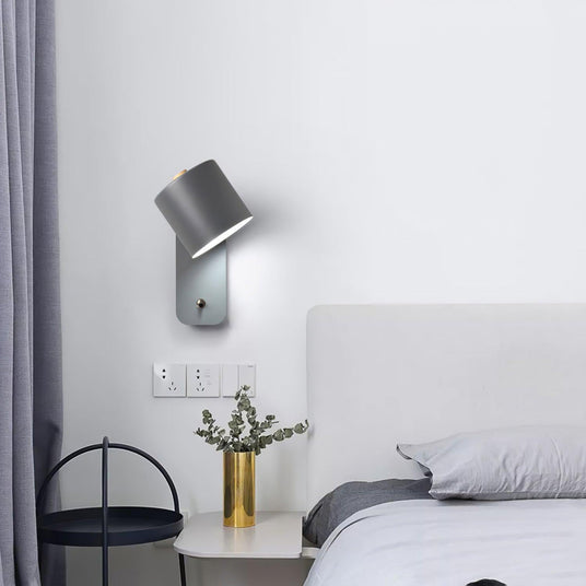 Lit above a bedside table with a brass vase, the bed and curtain just behind