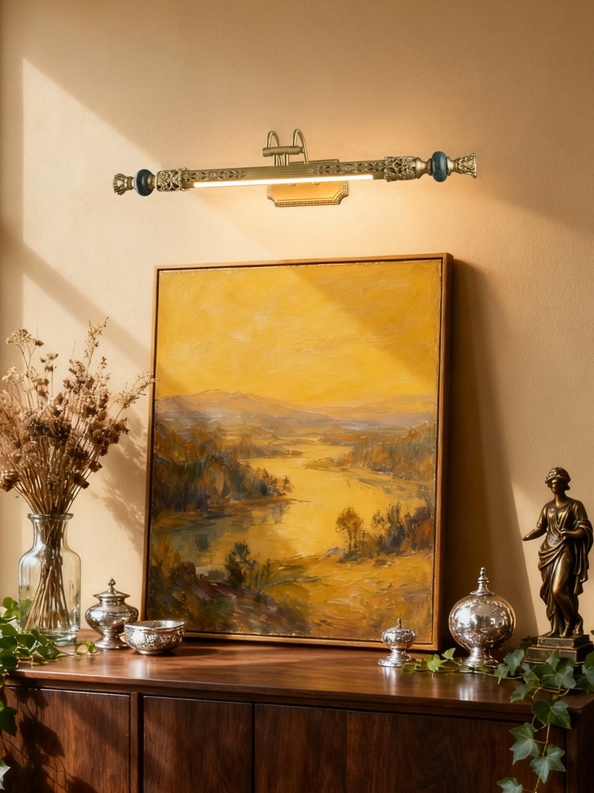 Ornate brass bar lit over a landscape painting above a dark wood chest