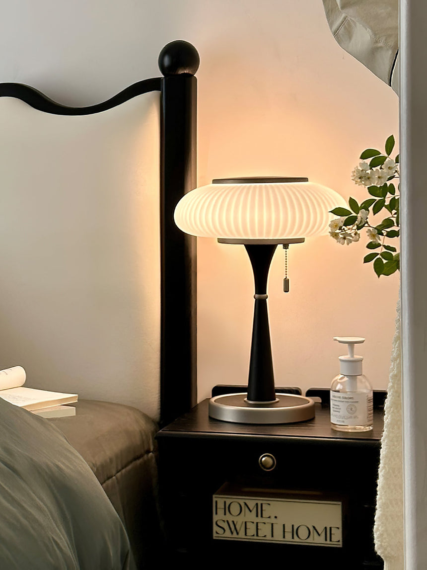 White glass table lamp lit on a nightstand beside a bed