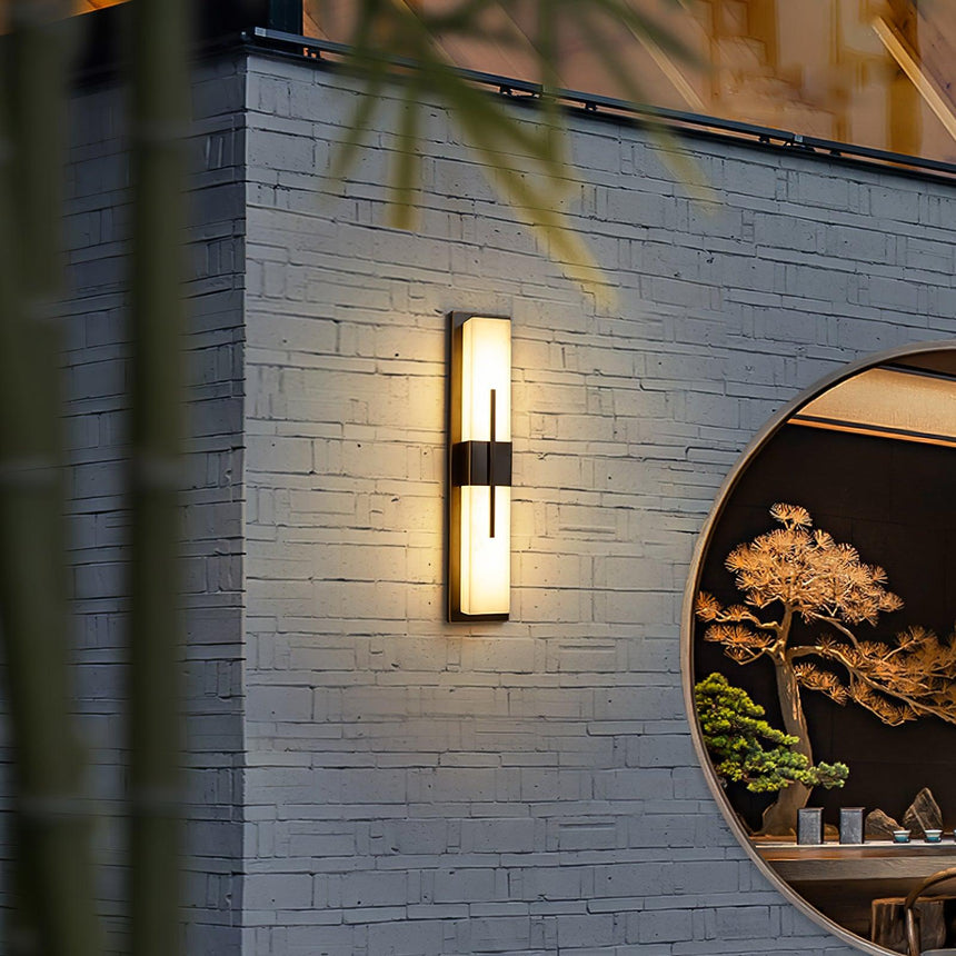 The light on a gray brick wall beside a round window framing a bonsai