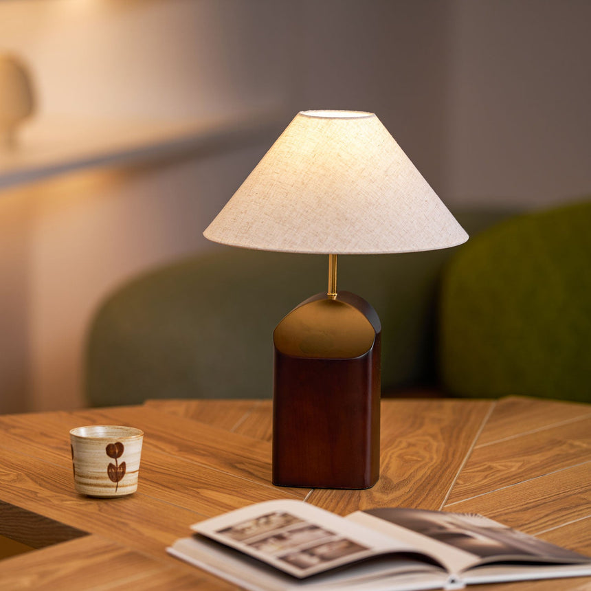 Bedroom table lamp lit on a wooden table beside a mug and a magazine