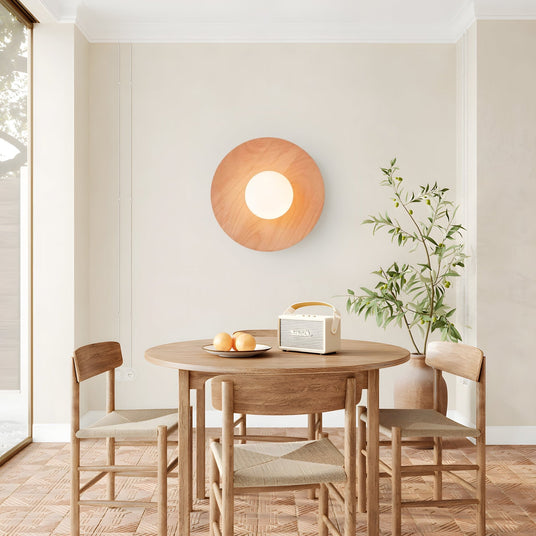 Lamp on a dining room wall beside a round table, chairs and an olive tree