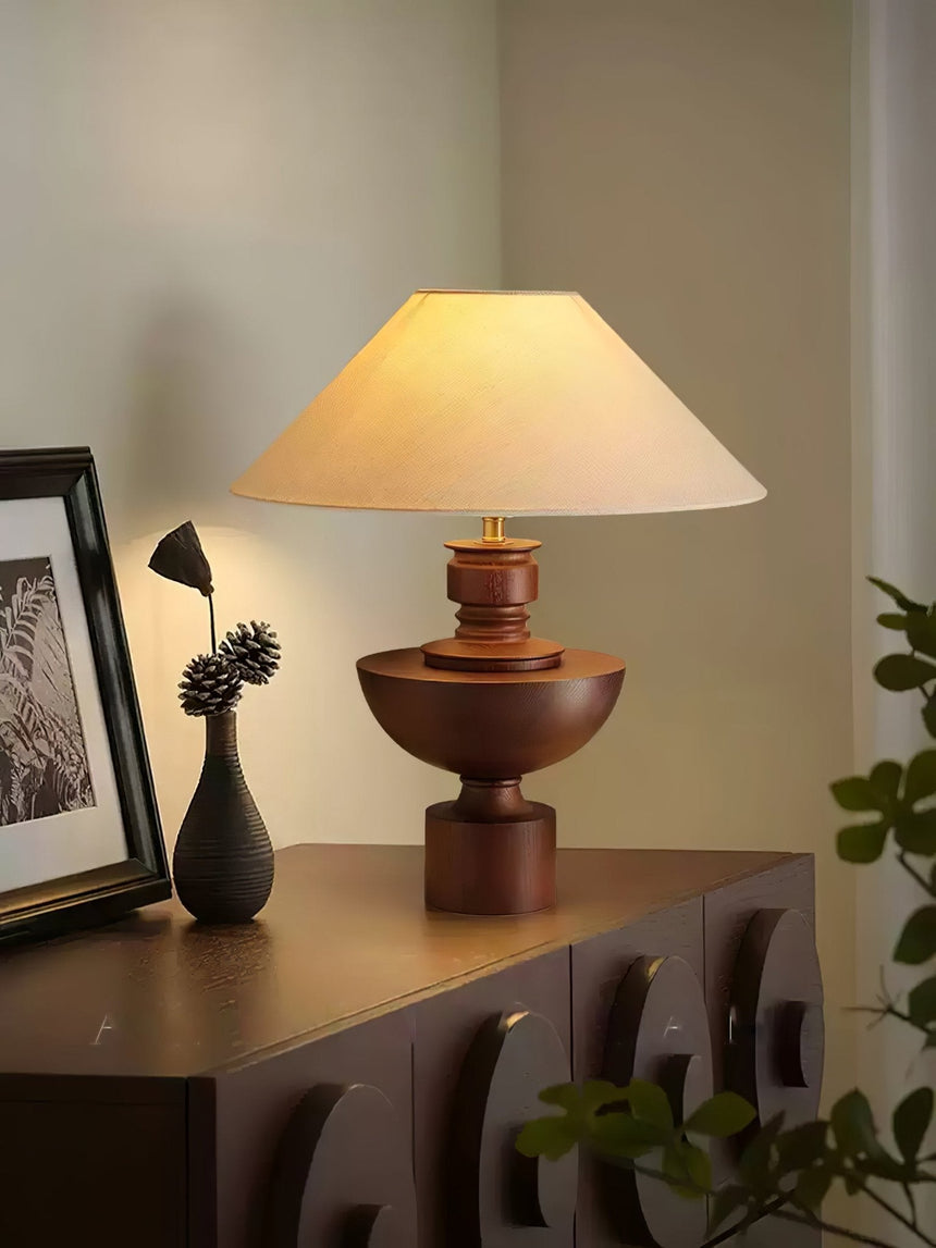 Beige table lamp lit on a dark sideboard beside a trailing plant