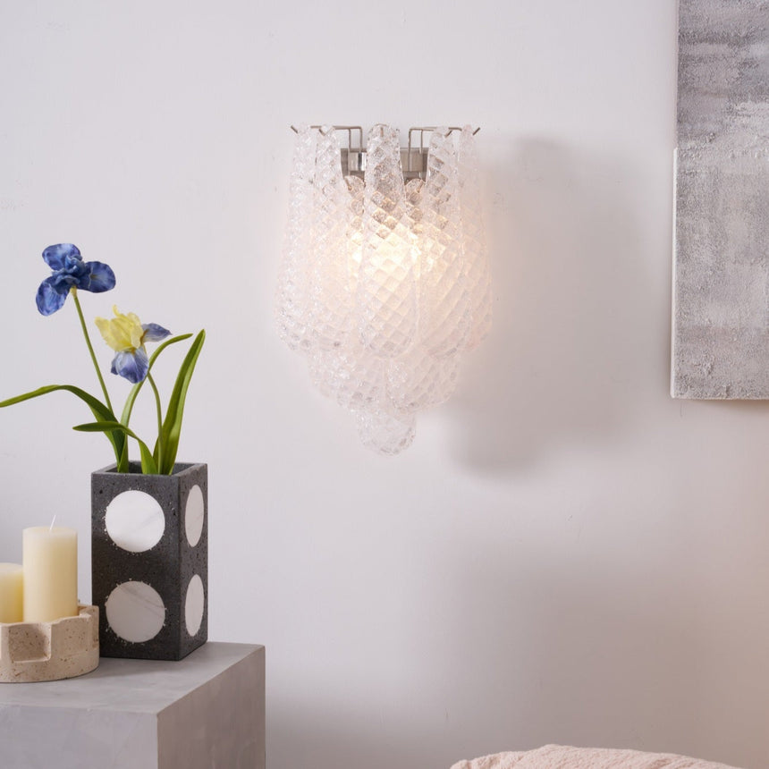 The clear sconce lit above a table with a vase of blue irises
