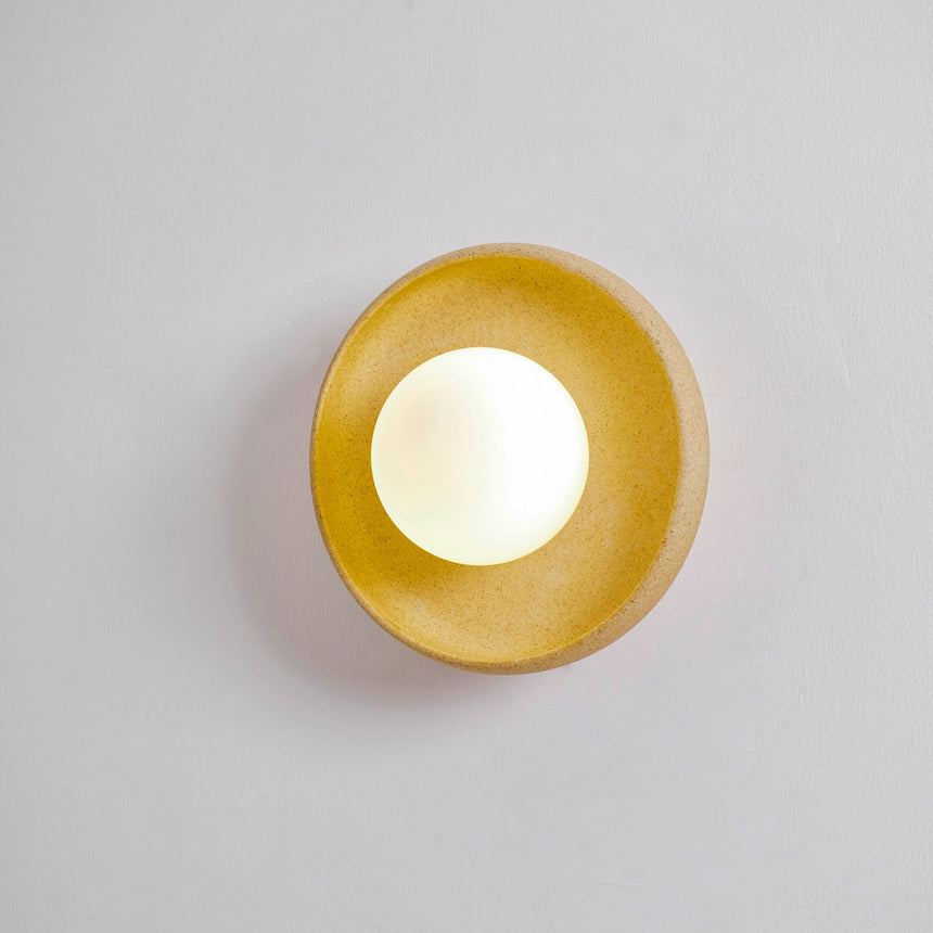 The round sconce lit on a pale wall, the glaze turning warm gold