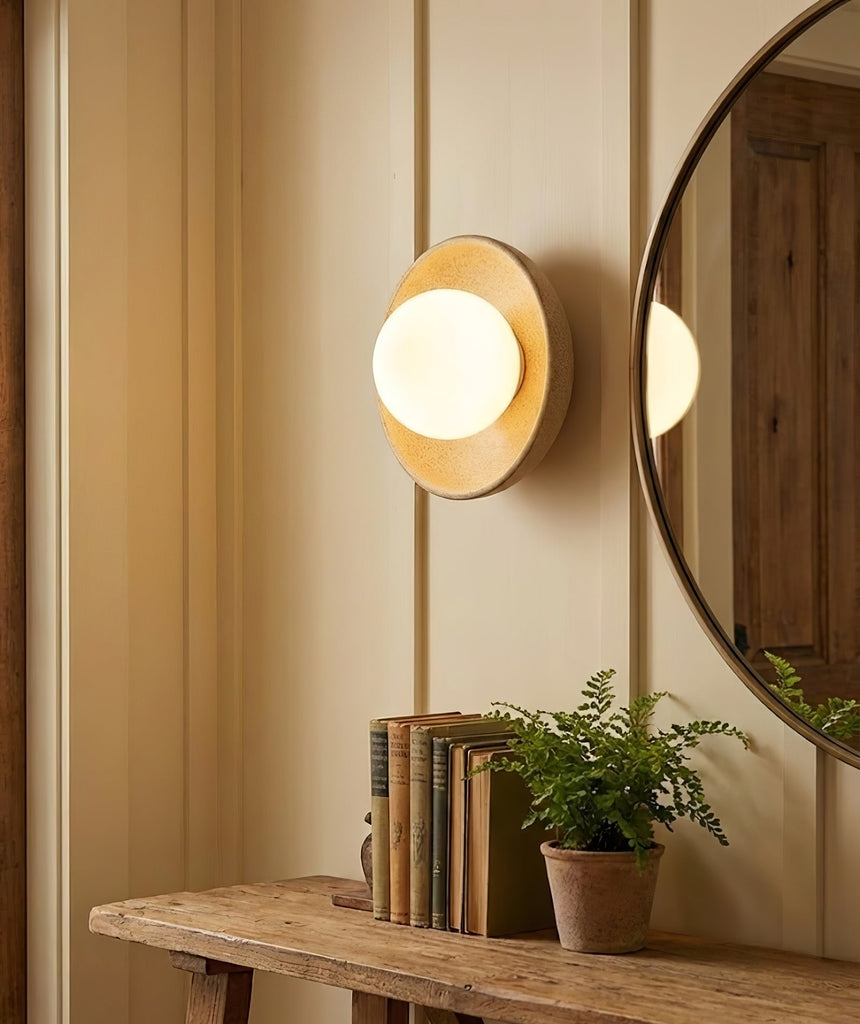 The sconce lit beside a round mirror above a wood console with books and a fern
