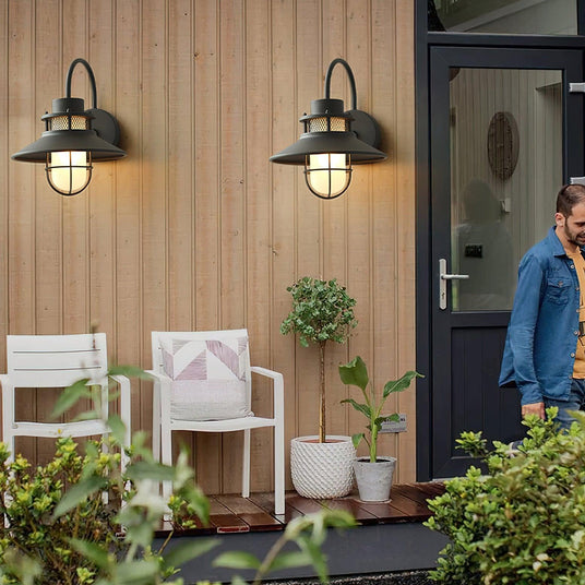 A pair of cage sconces lit on a wood clad wall beside a front door, white chairs on the porch