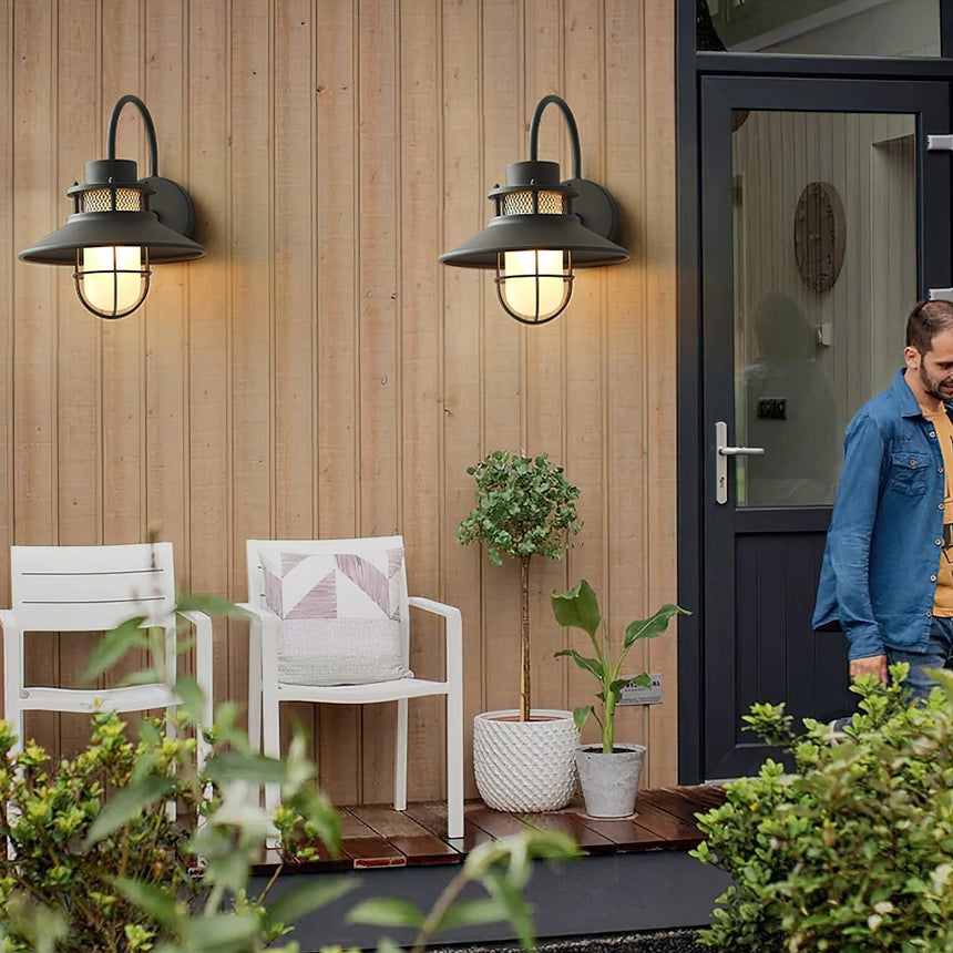 A pair of cage sconces lit on a wood clad wall beside a front door, white chairs on the porch