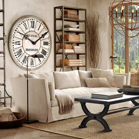 The lit lamp beside a pale sofa in a living room with a large wall clock and shelves
