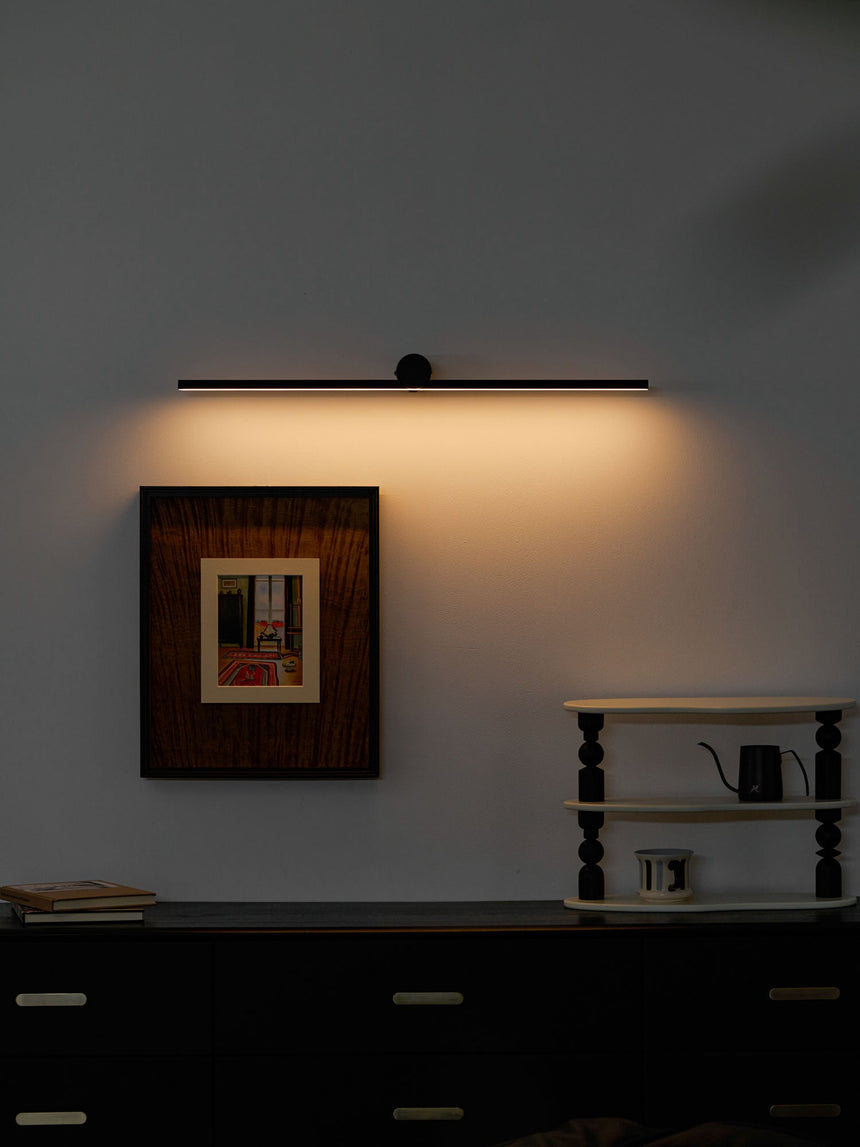 Black bar lit above a framed print on a dark wall, warm light washing down it