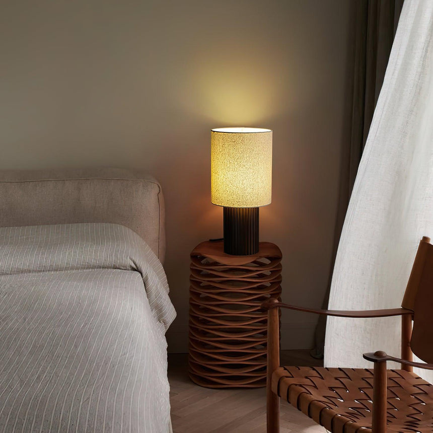 Entryway table lamp lit on a woven side table beside a bed and curtain