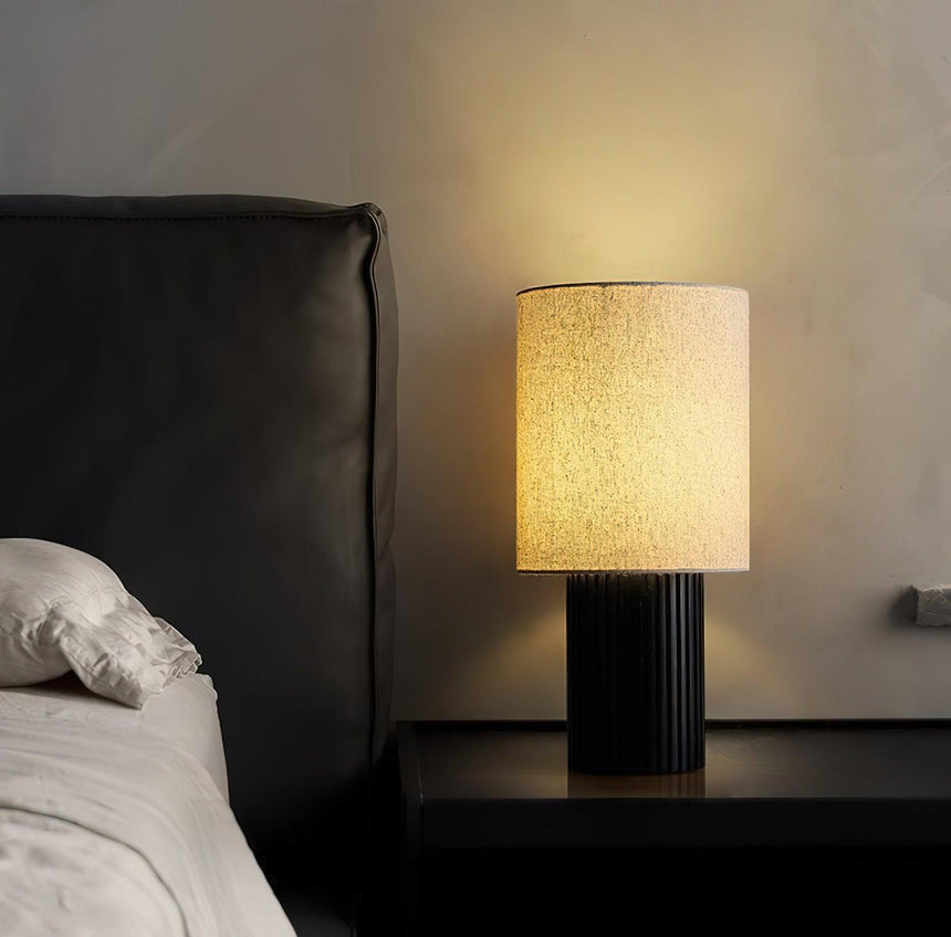 Entryway table lamp lit on a black nightstand beside a made bed