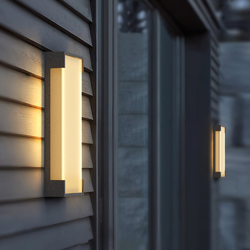 The sconce lit on gray lap siding beside a dark window reveal