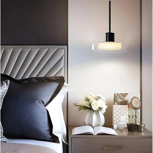 A glass disc pendant lit above a nightstand beside a tufted headboard and an open book