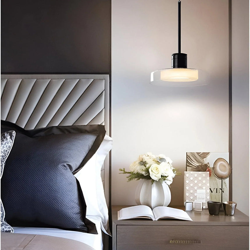 A glass disc pendant lit above a nightstand beside a tufted headboard and an open book
