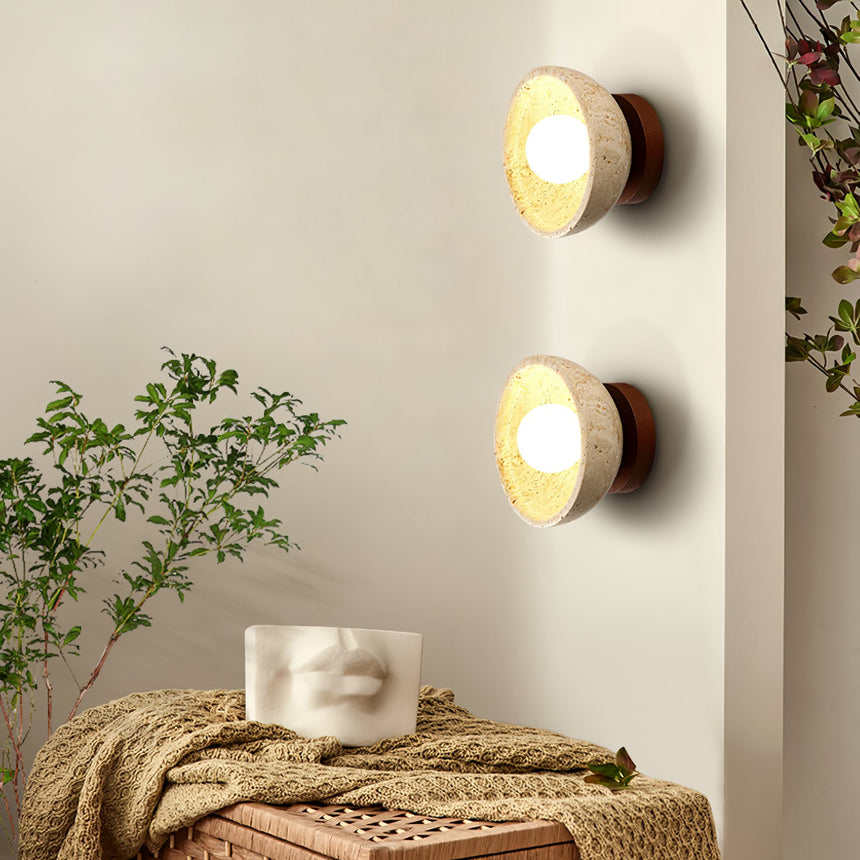 Two sconces lit beside a plant above a bench with a knit throw and a stone