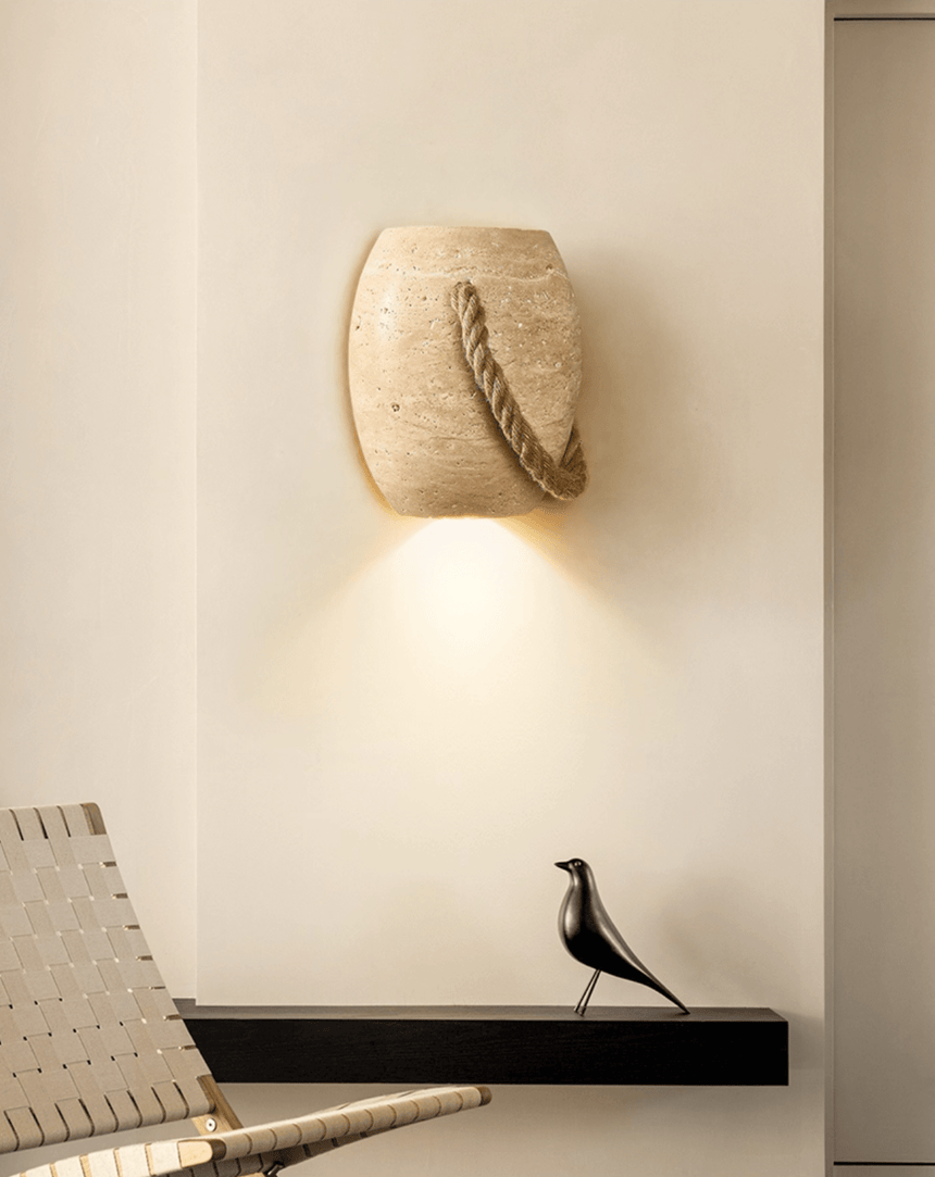 The lamp lit in the corner of a cream room above a black shelf and a woven chair back