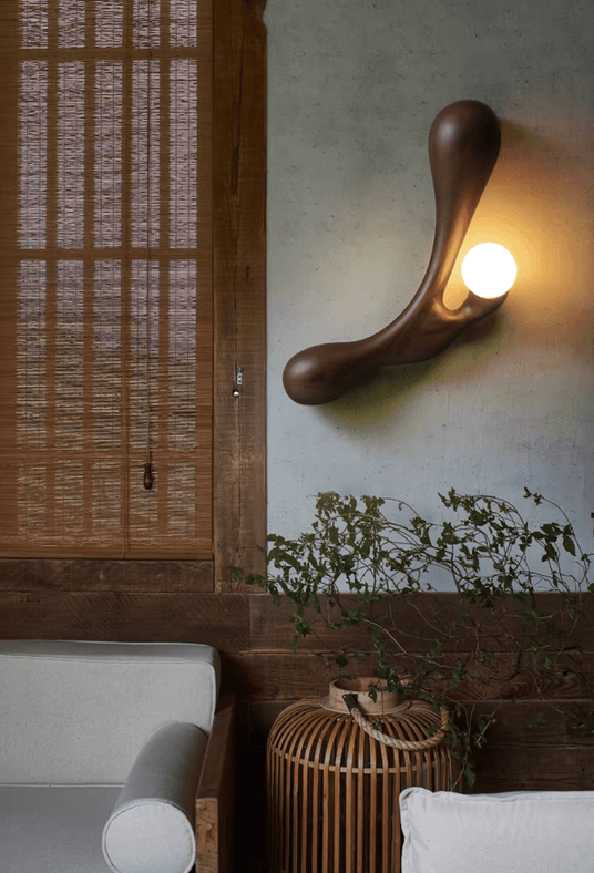 Sconce lit beside a slatted wood screen and a window with greenery beyond