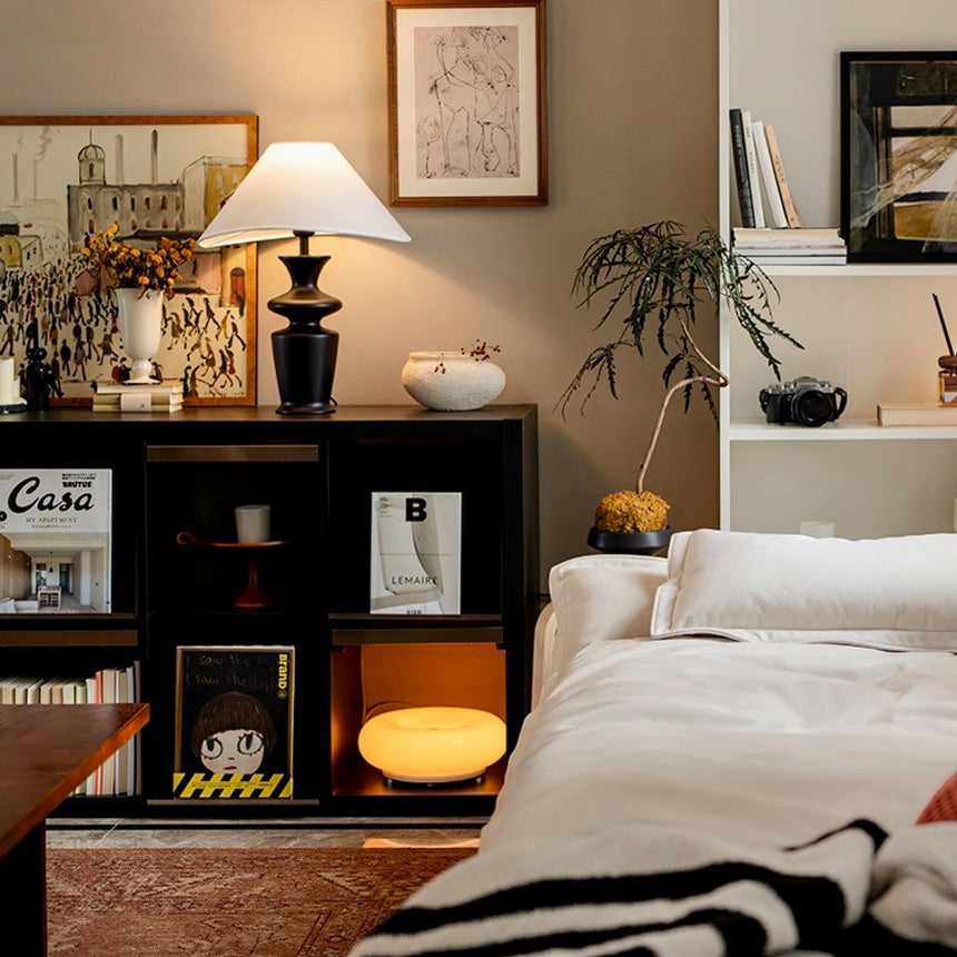 Large table lamp lit on a dark shelf unit beside a made bed