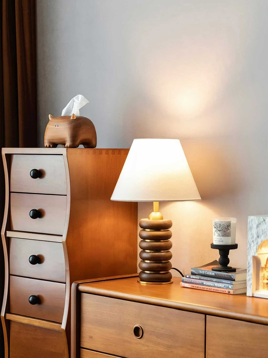 Linen shade table lamp lit on a wood dresser beside a drawer chest