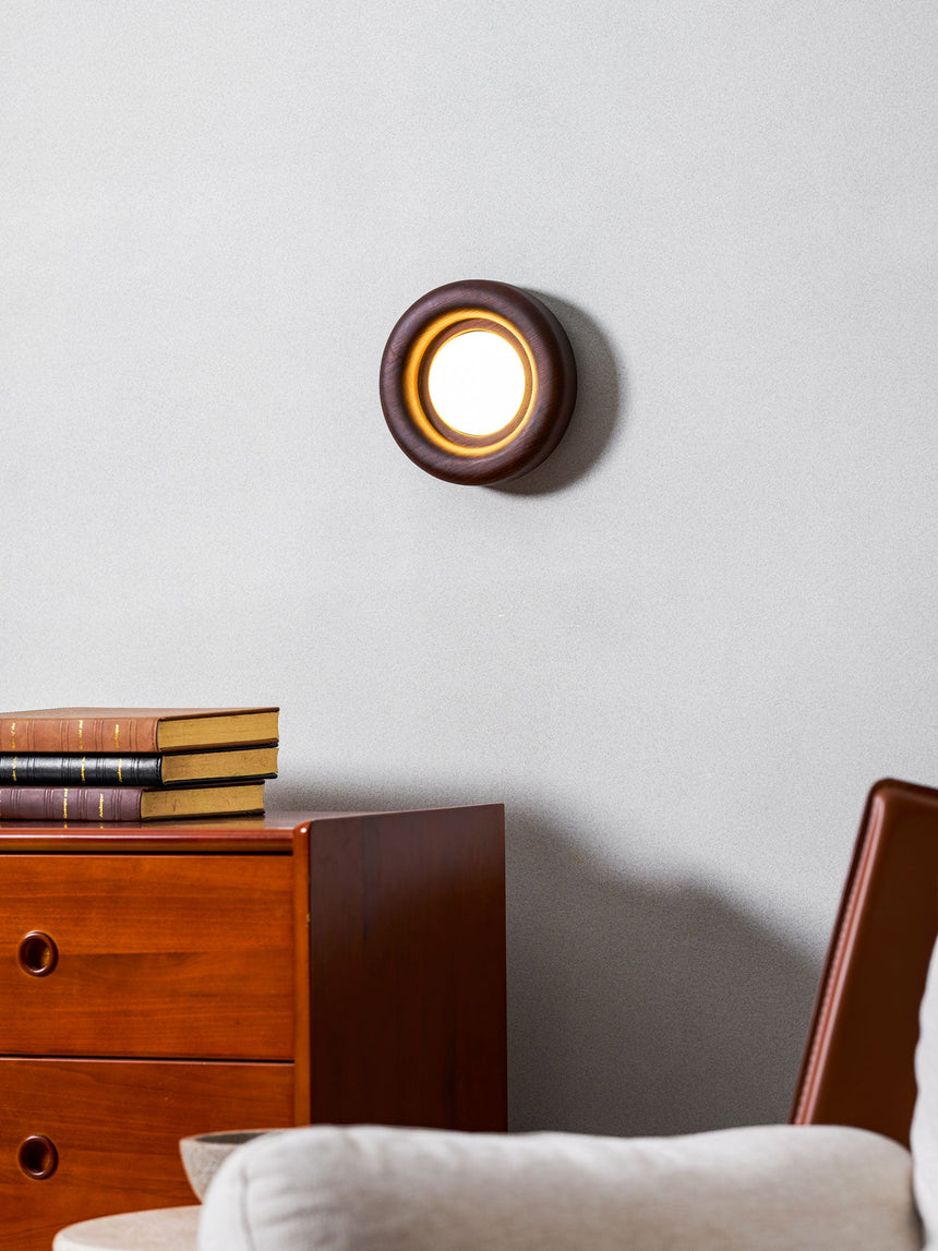 Sconce lit above a teak dresser beside a pale armchair with a cushion