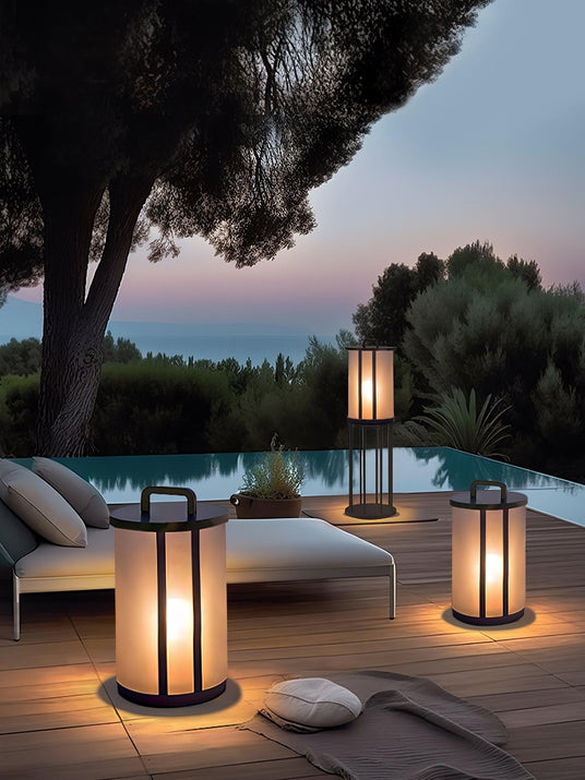 Two lanterns lit beside a pool at dusk with loungers and trees behind