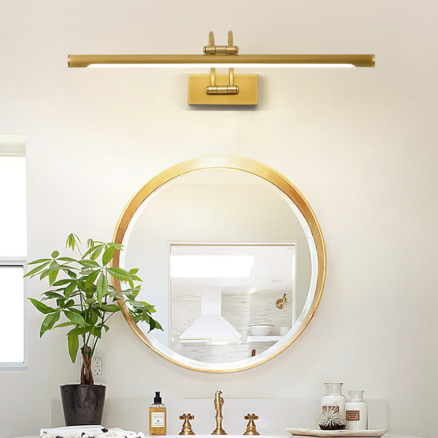 Brass bar lit above a round gold mirror on a vanity with a plant and glass bottles