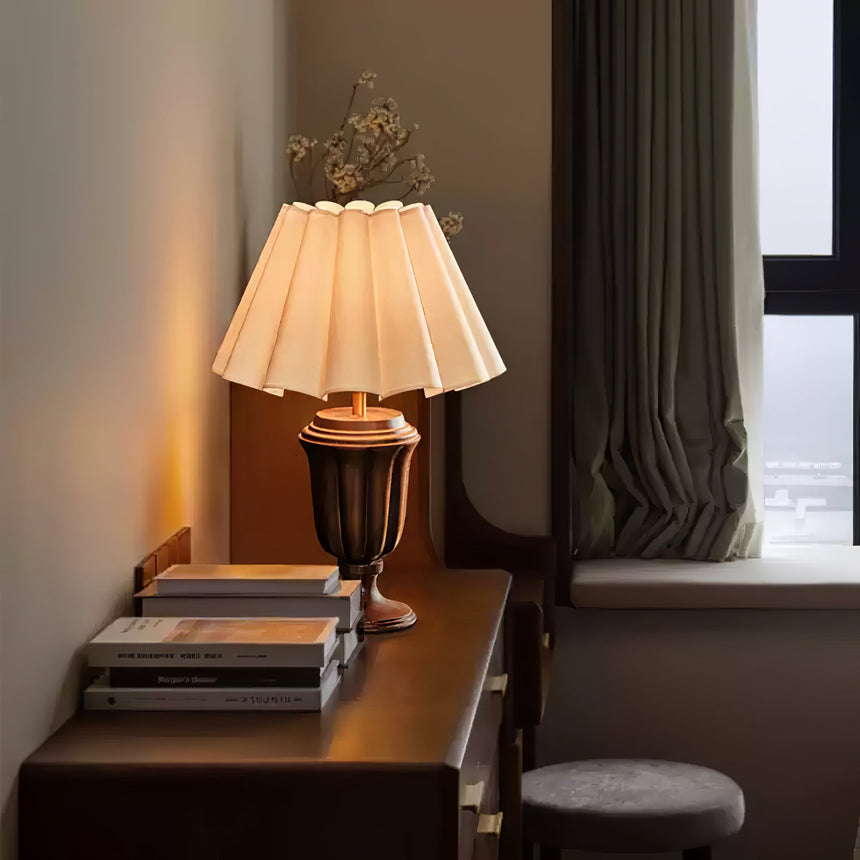 Urn base lamp lit on a dark desk beside stacked books and a stool