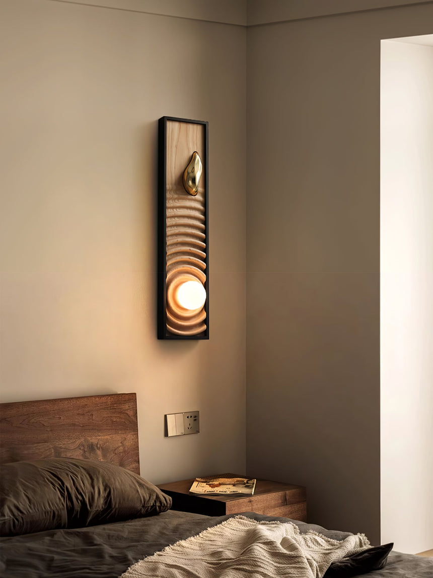 Sconce lit on a bedroom wall in a corner beside a bed with folded linen