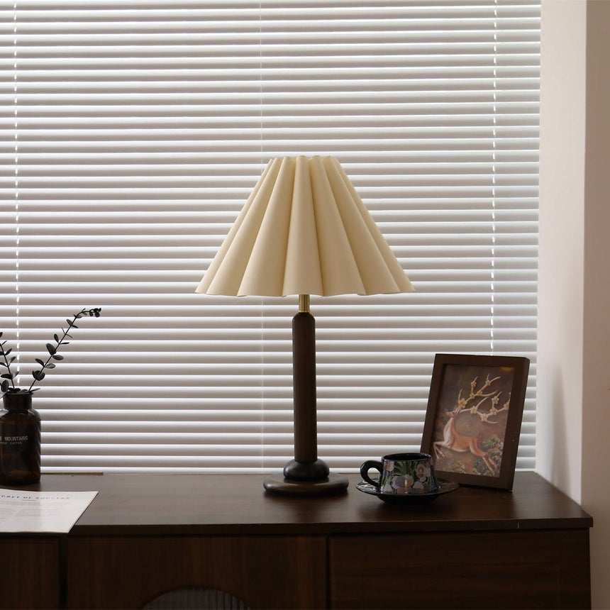 Pleated table lamp unlit on a dresser in front of window blinds