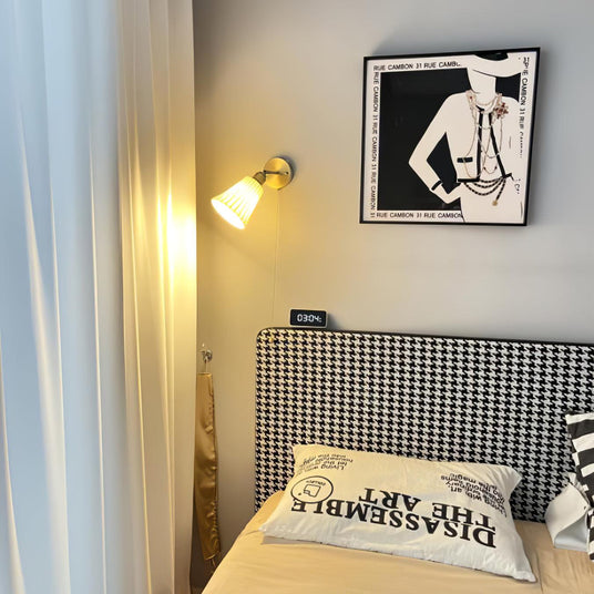 The sconce lit on a wall above a houndstooth headboard and a framed print