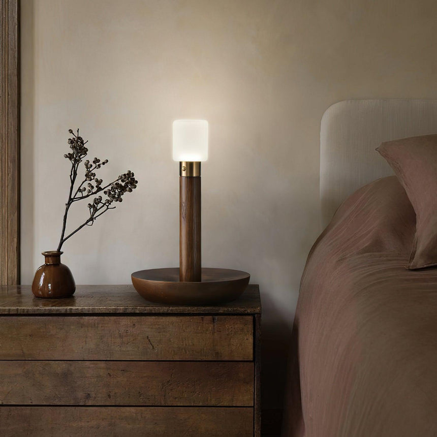 Portable table lamp lit on a wood dresser beside a small vase of twigs