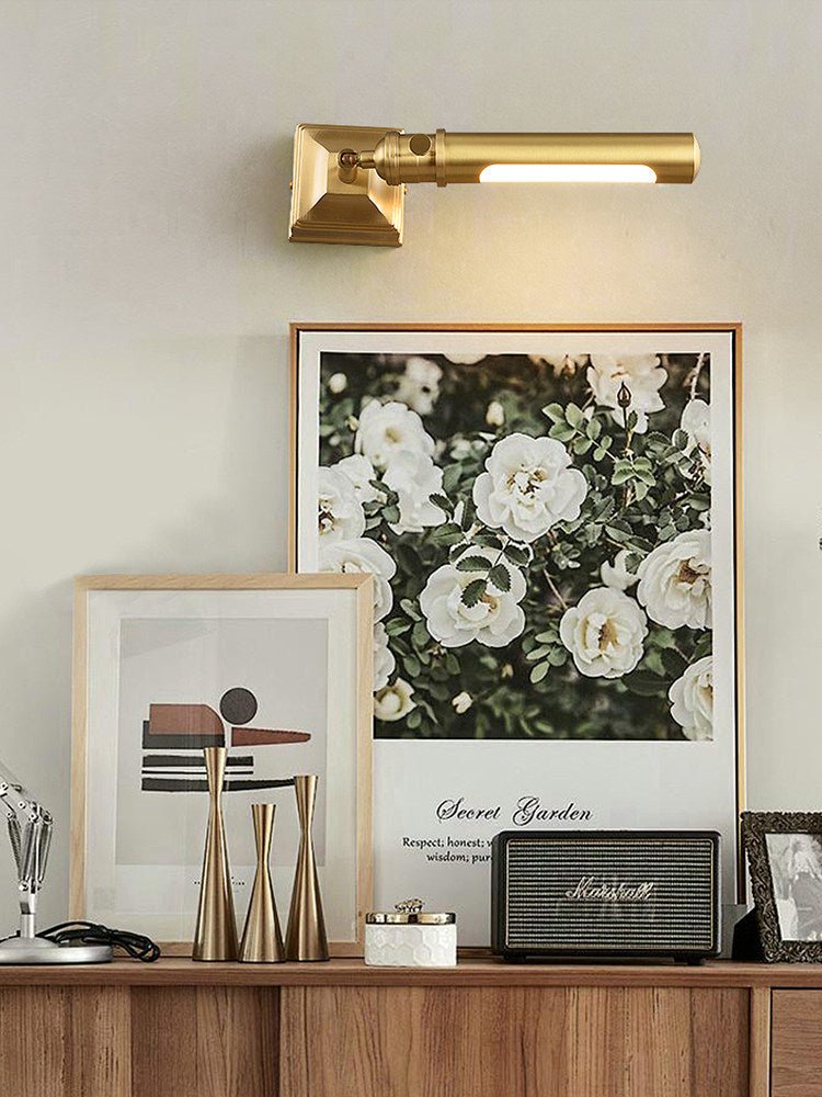 Brass tube lit above framed prints on a wood desk with pens and boxes