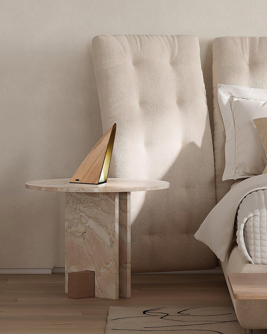 Small bedside lamp lit on a round stone table beside a tufted headboard