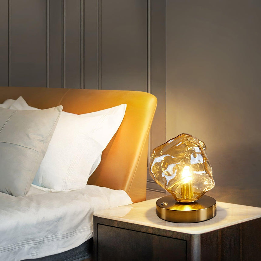 Glass ice form lamp lit on a white nightstand beside pillows
