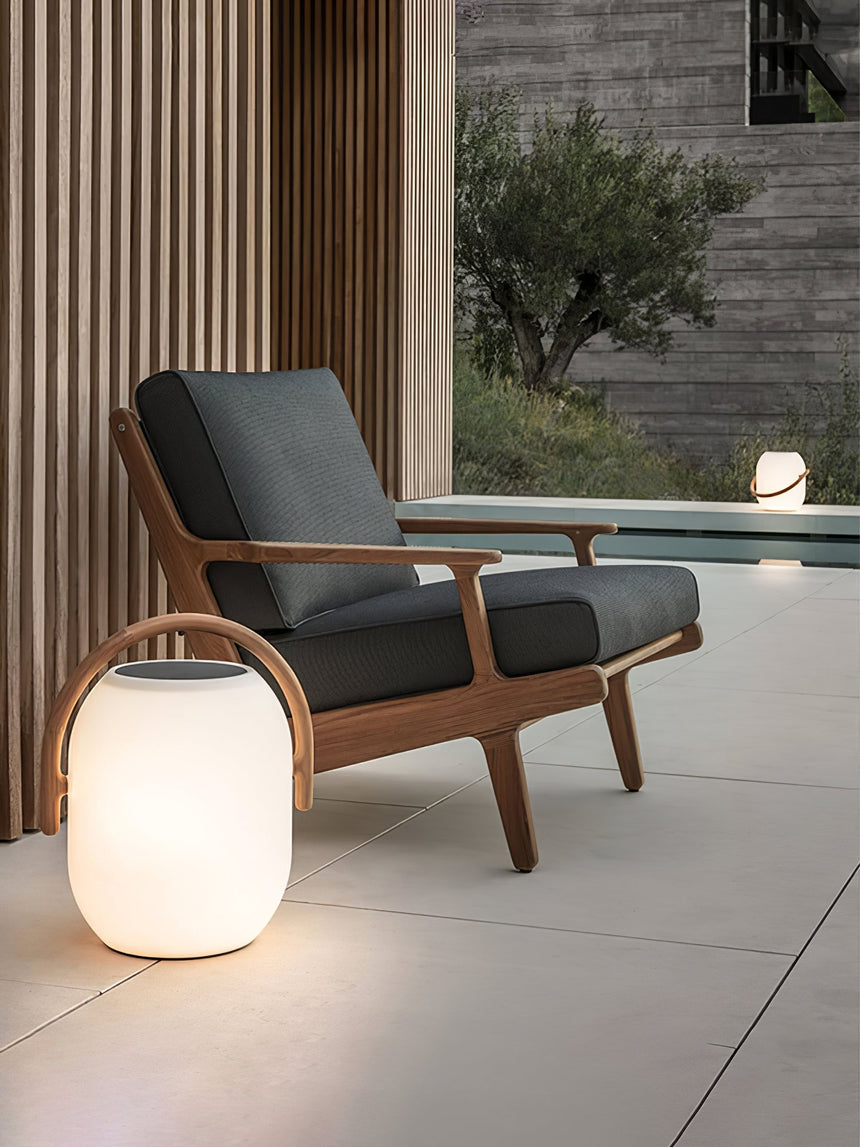 The lamp lit on paving beside a timber armchair at the edge of a pool