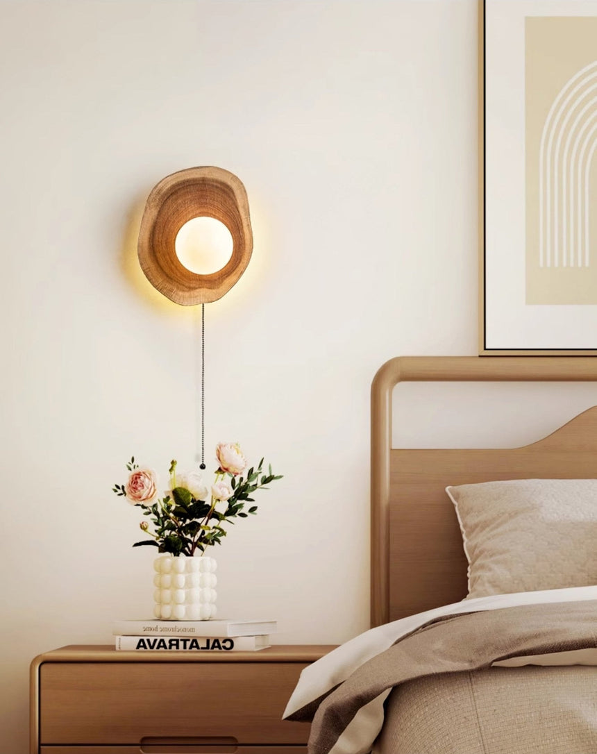 Sconce lit above a nightstand of flowers beside a bed with pale cushions