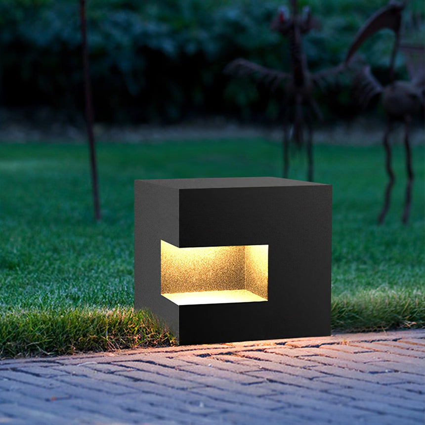 The cube lit low on paving beside grass, light spilling from the slot