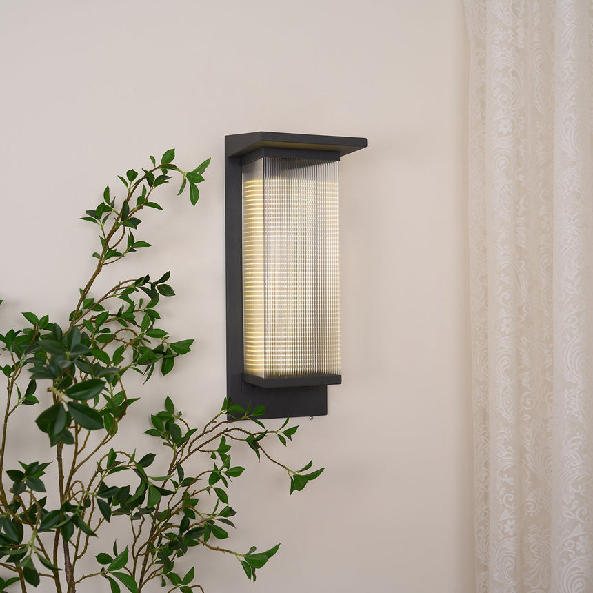 The box lantern lit on a pale wall beside a sheer curtain, a leafy plant in front