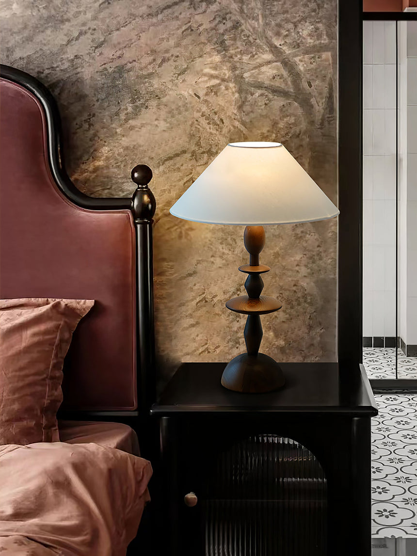 Spindle table lamp lit on a dark nightstand beside a bed with pink linen