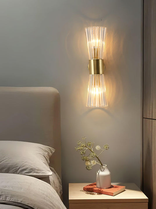 The sconce lit beside a gray headboard above a nightstand with a small vase