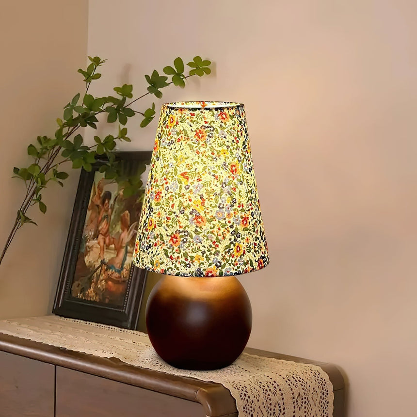 Floral shade lamp lit on a wood bench beside a framed botanical print