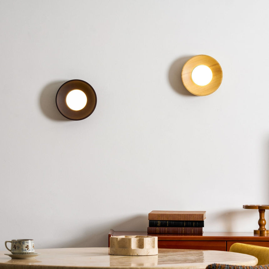 Two sconces on a wall above a desk with books, one walnut and one light wood