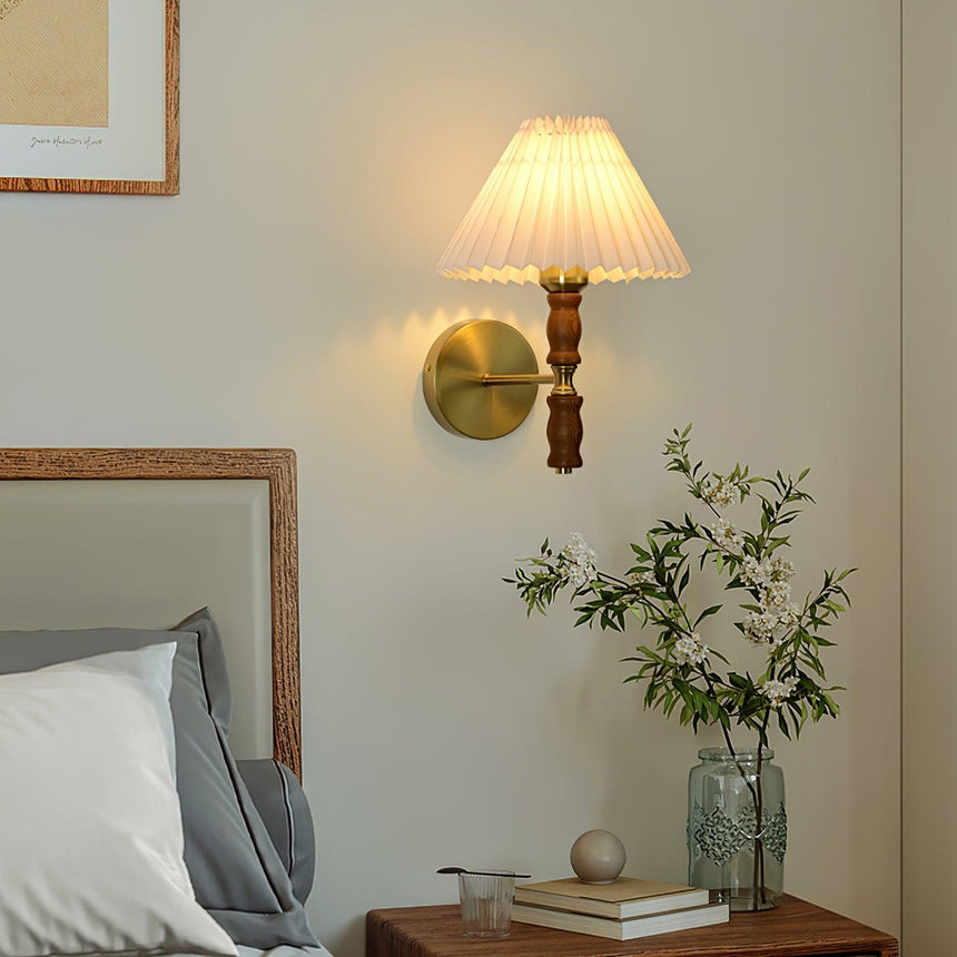 Pleated wall sconce lit above a nightstand with a vase of green stems