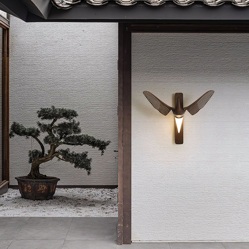 Sconce lit beside a doorway with a bonsai on a gravel bed beyond it