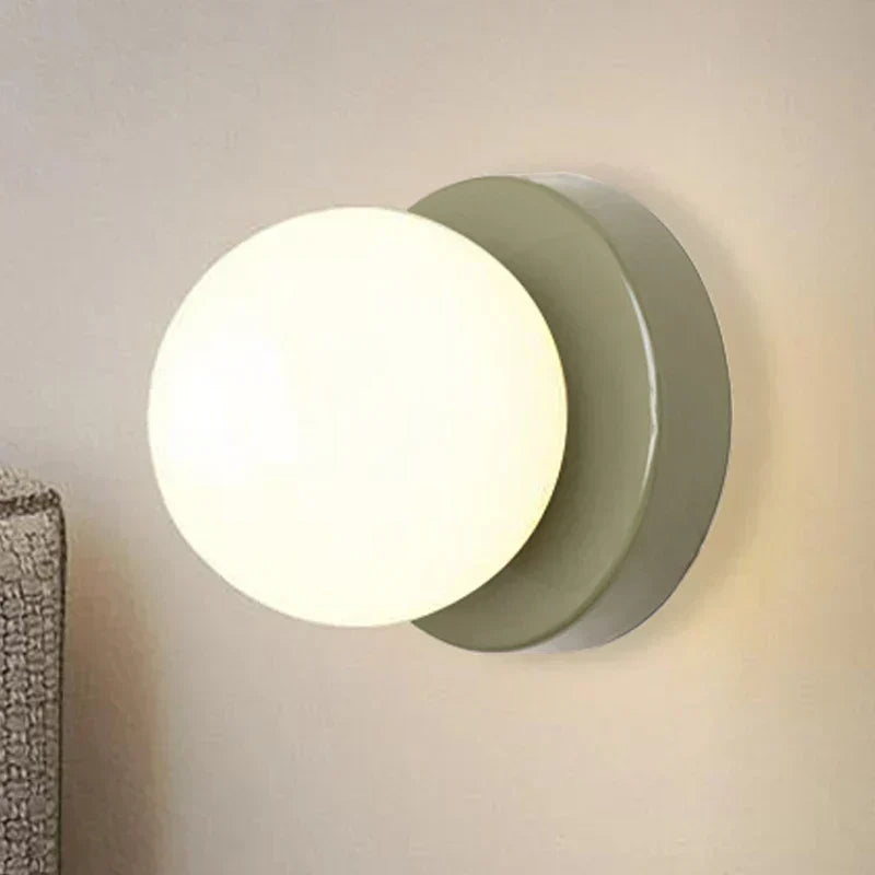 Iron glass globe wall sconce with slim round shade glowing warm white on a bedroom wall beside a soft neutral headboard
