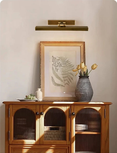 Brass bar lit over a framed print above a cane cabinet with yellow tulips