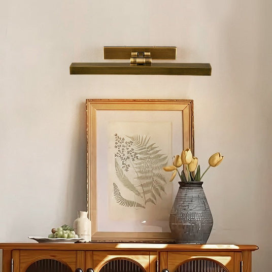 Brass bar lit above a framed fern print over a wood sideboard and a vase
