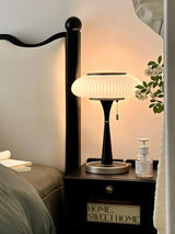 White glass table lamp lit on a nightstand beside a bed